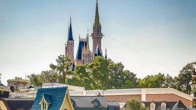 A castle with tall spires rises above treetops and rooftops, set against a clear blue sky. The scene includes buildings with blue and gray roofs in the foreground.