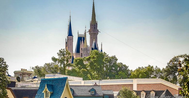 A castle with tall spires rises above treetops and rooftops, set against a clear blue sky. The scene includes buildings with blue and gray roofs in the foreground.