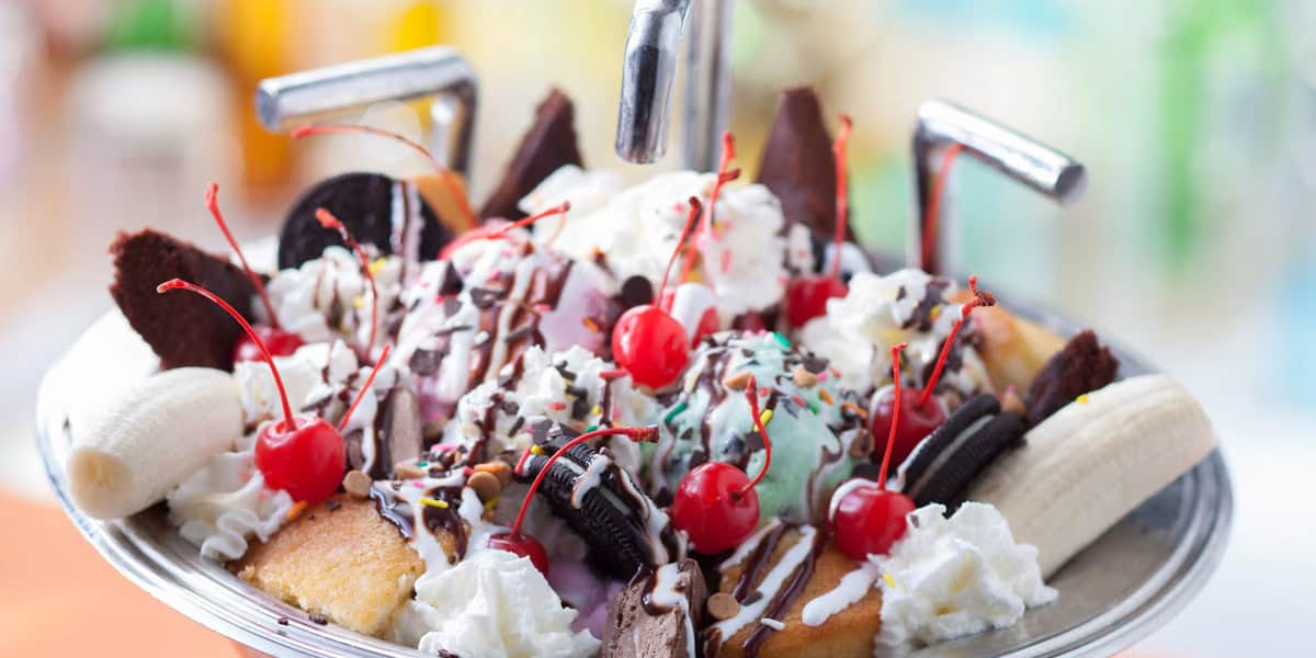 A large metal bowl filled with a banana split sundae, topped with scoops of ice cream, whipped cream, chocolate drizzle, sprinkles, cherries, brownies, cookies, and banana halves—just like a sweet treat from Disney Resort Activities—sits under two faucet-like handles.