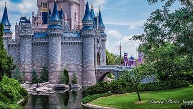 A fairytale castle with blue spires and grey stone walls stands beside a moat, surrounded by green trees and manicured lawns under a partly cloudy sky.