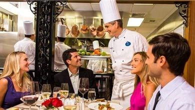 A chef in a white uniform and tall hat is smiling and speaking to four seated diners in a restaurant. The diners are dressed in semi-formal attire, sitting around a table with food and drinks. The kitchen is visible in the background through an open pass.