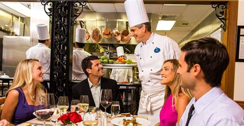 A chef in a white uniform and tall hat is smiling and speaking to four seated diners in a restaurant. The diners are dressed in semi-formal attire, sitting around a table with food and drinks. The kitchen is visible in the background through an open pass.