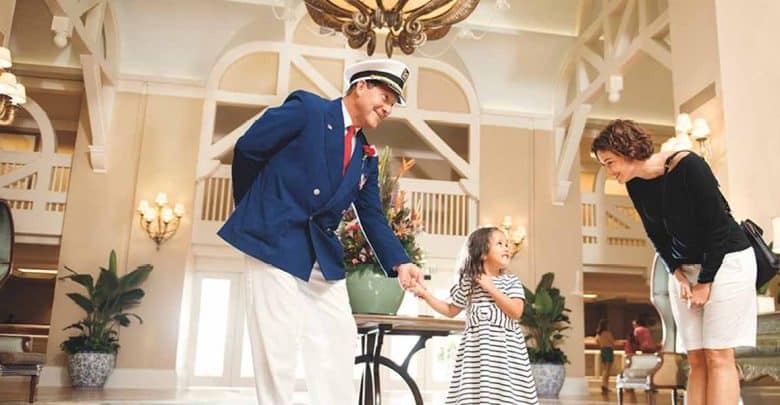 A hotel staff member in a blue blazer and white hat warmly greets a smiling young girl in a striped dress while her mother looks on, capturing the magic of luxury Disney experiences in a bright and elegant hotel lobby.