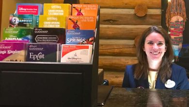 A friendly Disney Cast Member greets guests behind a desk with official park maps and brochures neatly arranged on display.