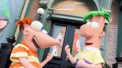 Two cartoon characters with large eyes and colorful hair, one with orange stripes and the other with green hair, stand smiling in front of a building, excited to explore the New Attractions at Walt Disney World 2025. Other characters appear through the windows behind them.