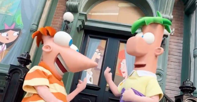 Two cartoon characters with large eyes and colorful hair, one with orange stripes and the other with green hair, stand smiling in front of a building, excited to explore the New Attractions at Walt Disney World 2025. Other characters appear through the windows behind them.