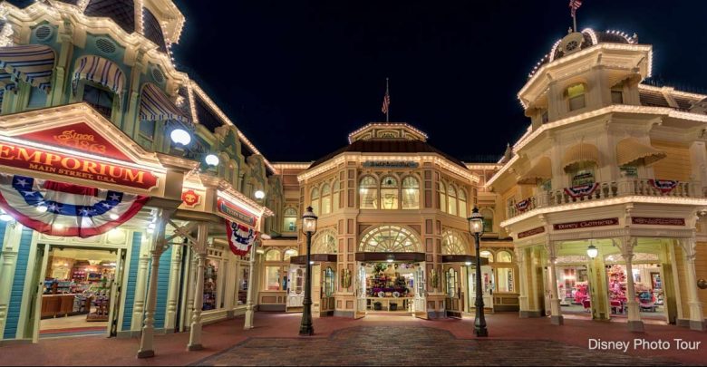 A nighttime view of a brightly lit Main Street scene features ornate buildings, including an Emporium with colorful banners. The area is illuminated, revealing hidden details in Magic Kingdom Park architecture and a welcoming entrance.