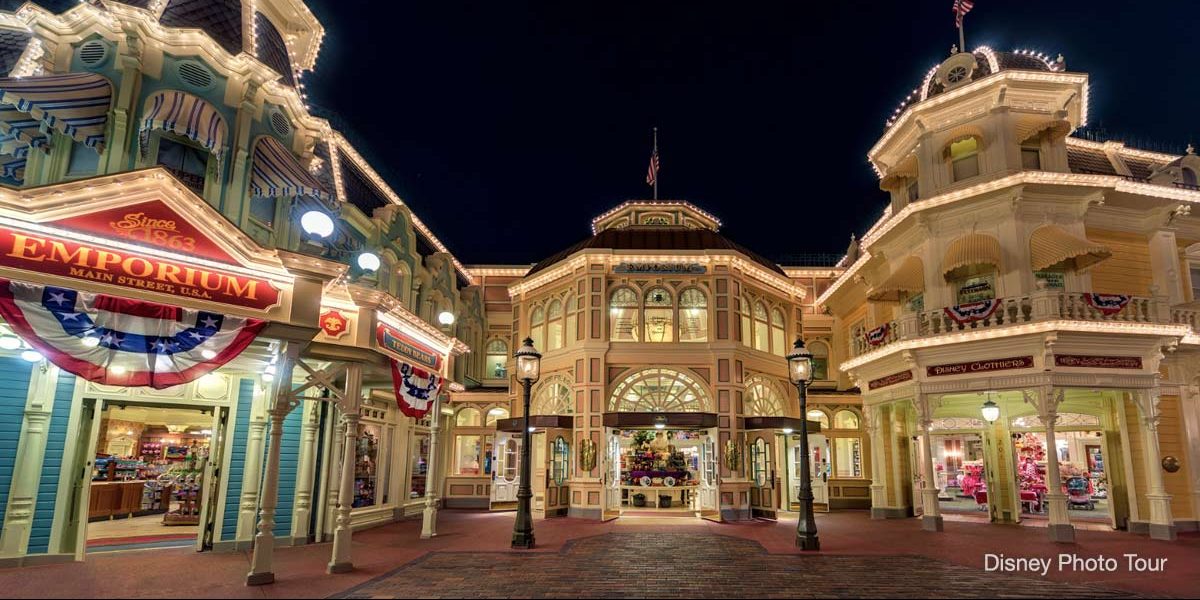 A nighttime view of a brightly lit Main Street scene features ornate buildings, including an Emporium with colorful banners. The area is illuminated, revealing hidden details in Magic Kingdom Park architecture and a welcoming entrance.