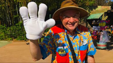 A smiling man in a safari hat and colorful shirt waves with a large white glove, standing outdoors near a souvenir stand with plush animals and hats.