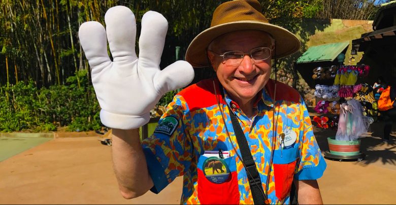 A smiling man in a safari hat and colorful shirt waves with a large white glove, standing outdoors near a souvenir stand with plush animals and hats.