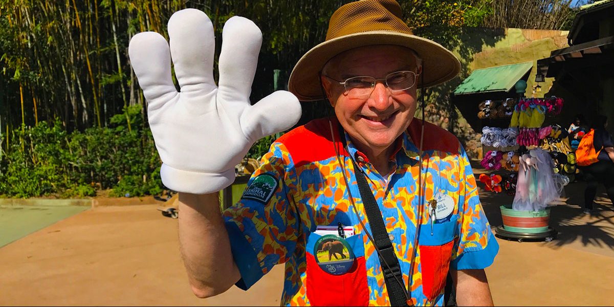 A smiling man in a safari hat and colorful shirt waves with a large white glove, standing outdoors near a souvenir stand with plush animals and hats.