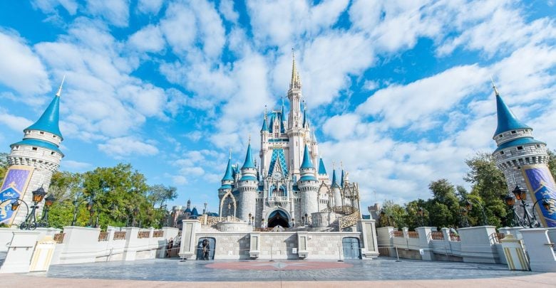 Cinderella Castle at a Disney theme park, photographed from the front under a blue sky with scattered clouds. The castle features tall spires and ornate towers, with trees and lamp posts on either side.