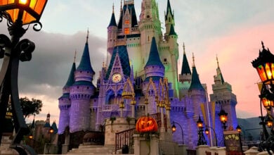 Cinderella Castle at Disney World shines with purple lights at sunset, surrounded by Halloween pumpkins and glowing lanterns.