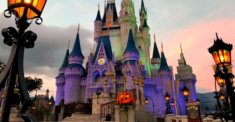 Cinderella Castle at Disney World shines with purple lights at sunset, surrounded by Halloween pumpkins and glowing lanterns.
