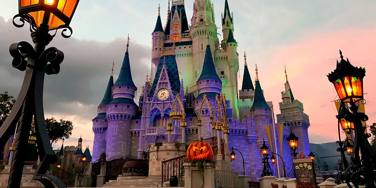 Cinderella Castle at Disney World shines with purple lights at sunset, surrounded by Halloween pumpkins and glowing lanterns.