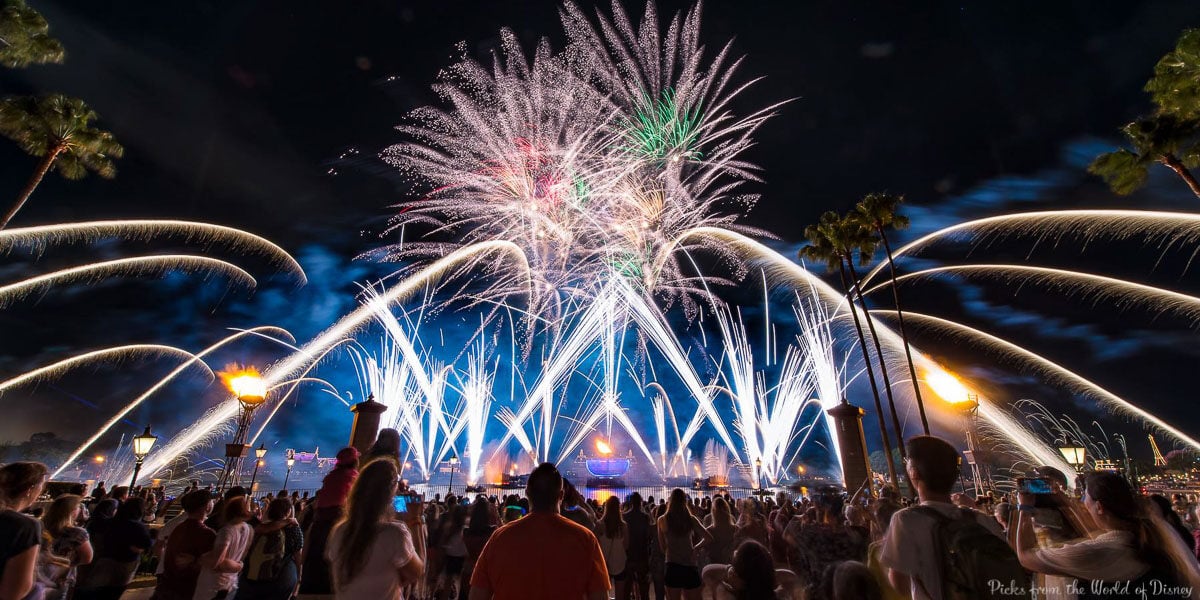 A crowd watches colorful fireworks and light streams fill the night sky above EPCOT's waterfront, surrounded by palm trees and glowing street lamps.