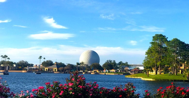 A scenic view of a lake with vibrant pink flowers in the foreground, trees along the shore, and a large white geodesic dome in the distance under a blue sky.