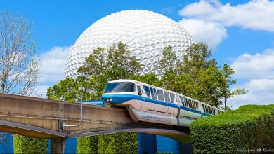 A Disney monorail glides past Spaceship Earth at Epcot, set against a bright blue sky dotted with fluffy clouds.