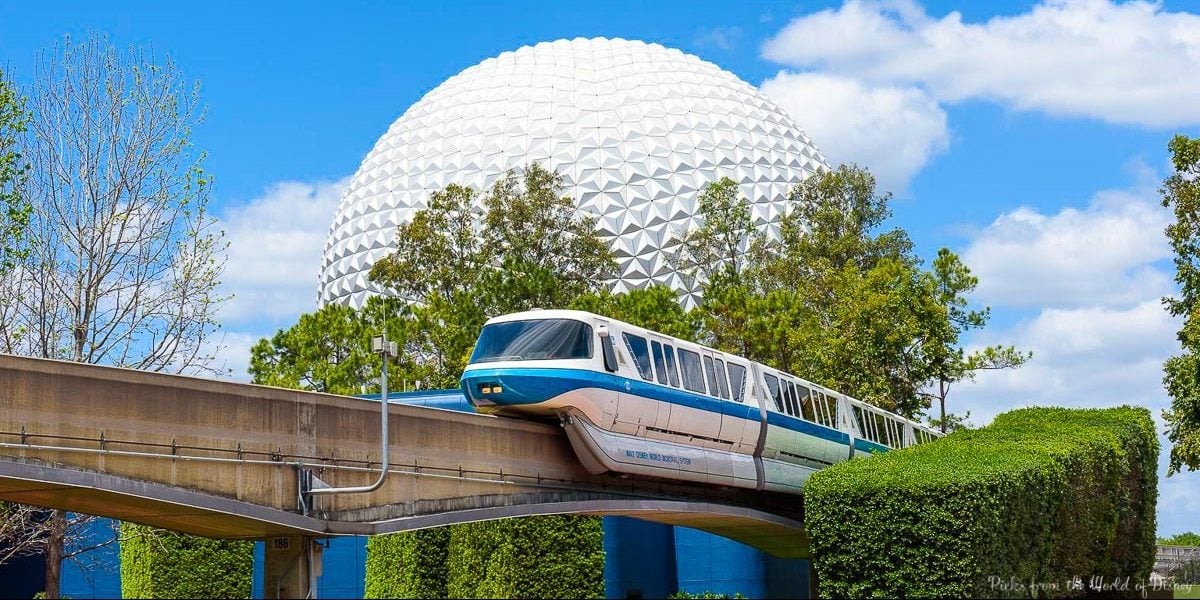 A Disney monorail glides past Spaceship Earth at Epcot, set against a bright blue sky dotted with fluffy clouds.