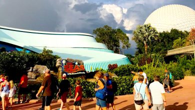 People walking near "The Seas with Nemo & Friends" attraction at Epcot, Disney World. The scene includes a large white geodesic sphere in the background and lush greenery, with a partly cloudy sky above.