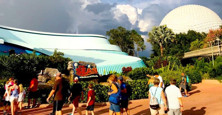 People walking near "The Seas with Nemo & Friends" attraction at Epcot, Disney World. The scene includes a large white geodesic sphere in the background and lush greenery, with a partly cloudy sky above.