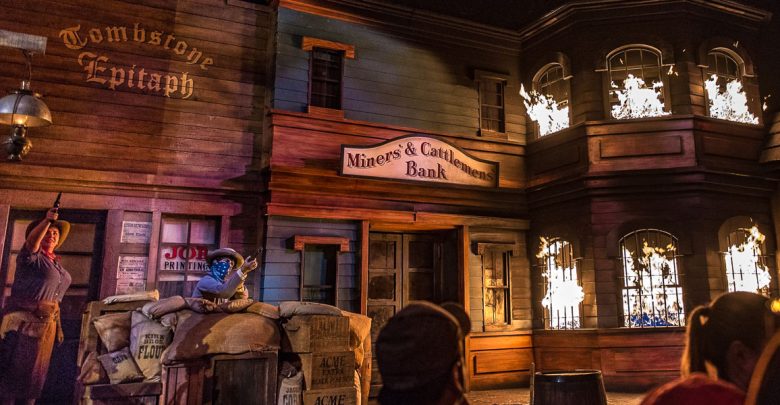 A western-themed scene with a bank and newspaper office, sacks and crates stacked outside, a person in a red bandana aiming a gun, and flames coming from the upper windows of the wooden buildings.