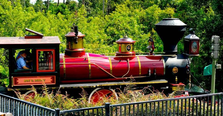 The Roy O. Disney steam train chugs along vibrant tracks at Walt Disney World, surrounded by lush green scenery under sunny skies.