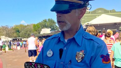 A security guard in a blue uniform and cap stands outdoors, holding a device. People walk behind him in a sunny, busy area with trees and buildings in the background.