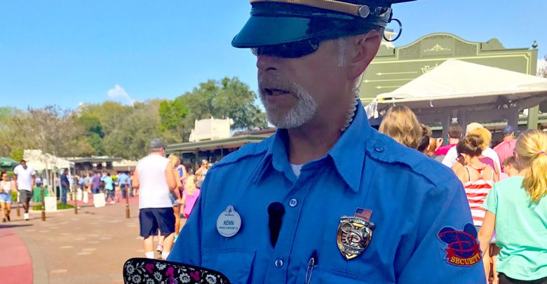 A security guard in a blue uniform and cap stands outdoors, holding a device. People walk behind him in a sunny, busy area with trees and buildings in the background.
