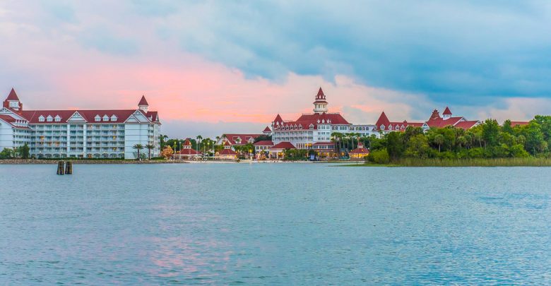 A large lake with a shoreline featuring white buildings topped with red roofs and turrets, surrounded by green trees under a cloudy sky at sunset, reminiscent of the Magic Kingdom Fireworks sparkling in the distance.