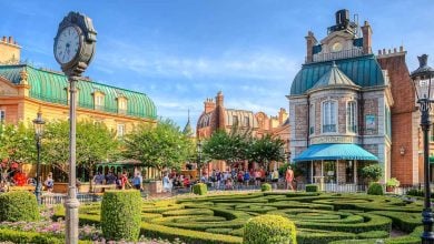 A lively square with a manicured garden maze, ornate street lamps, a large clock, and elegant European-style buildings; people stroll and gather on a sunny day under a bright blue sky.