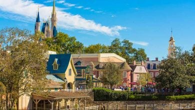 A colorful, bustling area of a theme park with colonial-style buildings, lush trees, and Cinderella Castle rising in the background under a bright blue sky—perfect for exploring the best attractions at Magic Kingdom in the morning.