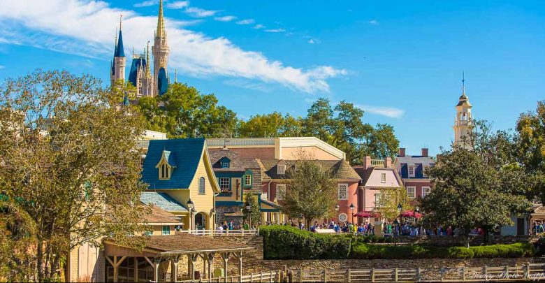 A colorful, bustling area of a theme park with colonial-style buildings, lush trees, and Cinderella Castle rising in the background under a bright blue sky—perfect for exploring the best attractions at Magic Kingdom in the morning.