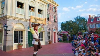Colonial Williamsburg performer lifts "Applause!" sign for the crowd during an outdoor show near historic buildings.