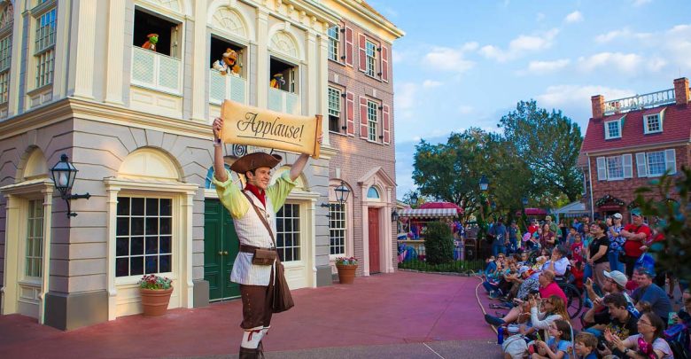 Colonial Williamsburg performer lifts "Applause!" sign for the crowd during an outdoor show near historic buildings.