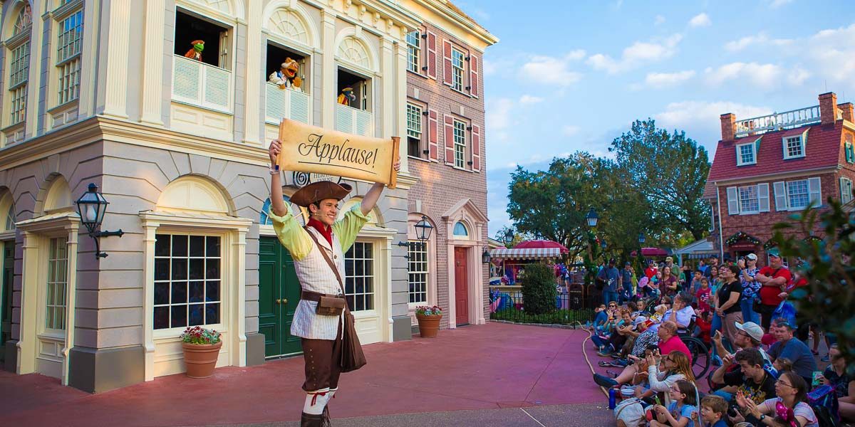 Colonial Williamsburg performer lifts "Applause!" sign for the crowd during an outdoor show near historic buildings.