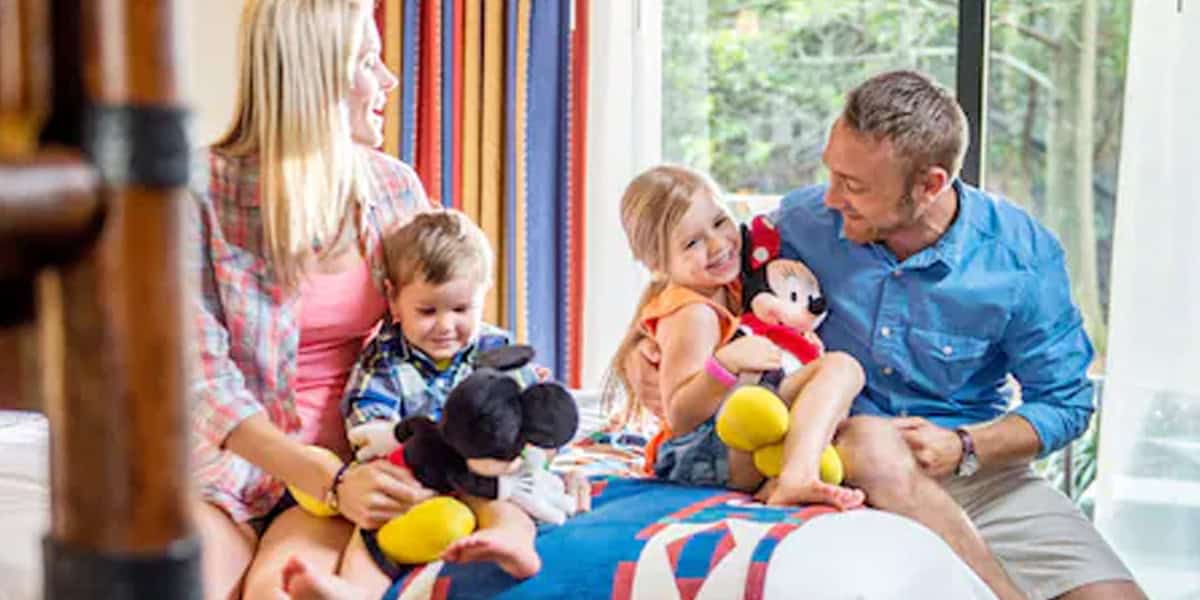 A family of four sits on a bed, smiling and holding Mickey and Minnie Mouse plush toys. Sunlight streams in through a large window, colorful curtains decorate the room, making it feel like a page from Walt Disney World Resort Tips.