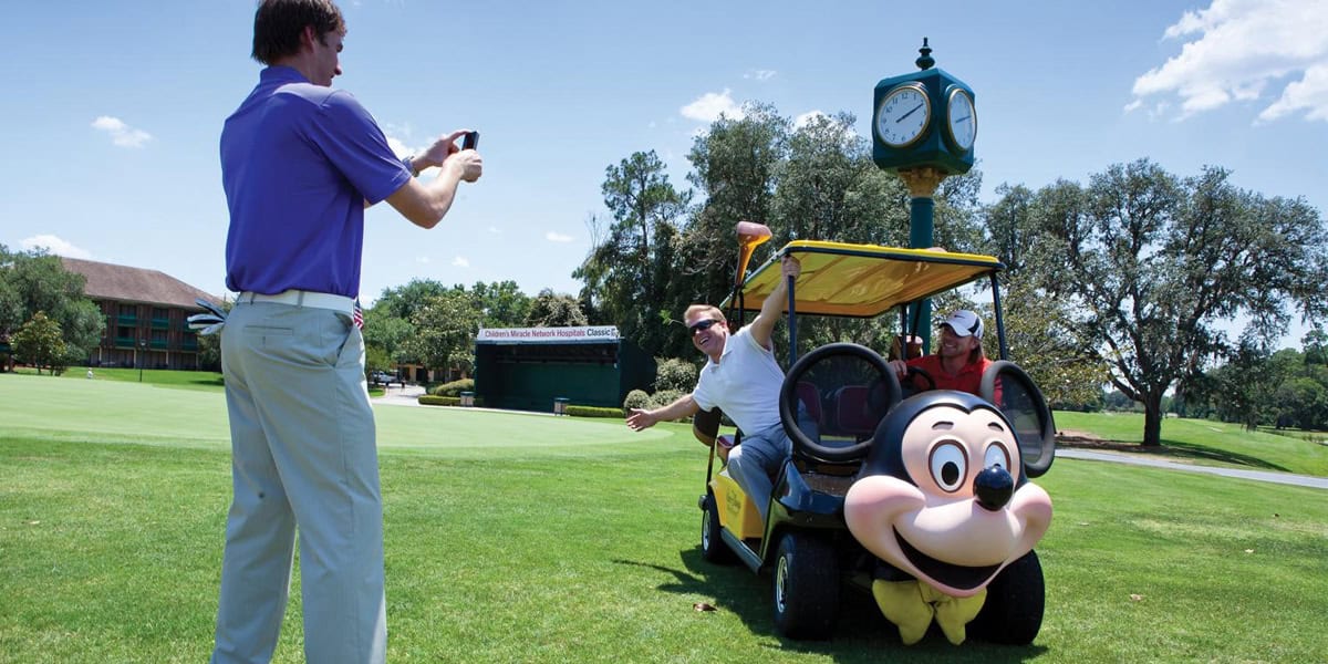 A man snaps a photo of two people posing in a golf cart decorated with a large Mickey Mouse face on a green golf course under a blue sky, with a clock tower and trees in the background—perfect for your Walt Disney World Resort tips album.