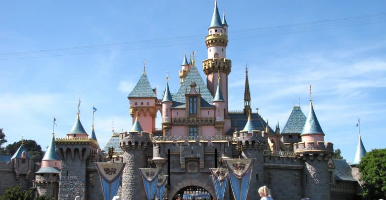 A view of a fairytale castle with blue and pink spires under a clear blue sky. Many people are gathered in front, taking photos and enjoying the scene. The castle is surrounded by trees and decorative flags.