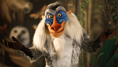 A costumed character with a colorful face, long white hair, and a grey furry body stands with arms open. The background is blurred with hints of greenery and animals.