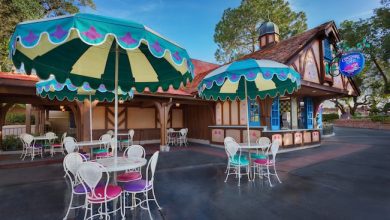 Charming cottage café with vibrant umbrellas and white chairs, perfect for relaxing between attractions at a lively theme park.