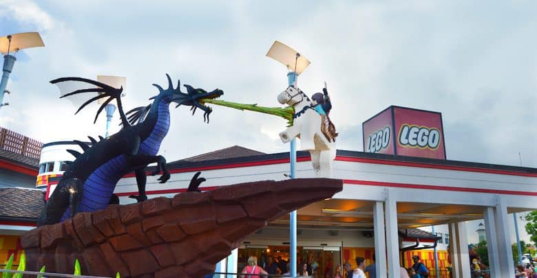 A Lego dragon faces off with a Lego knight on horseback outside the entrance to LEGOLAND, beneath dramatic cloudy skies.