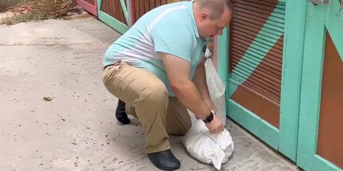 A man in khaki pants and a light blue shirt places a sandbag against a closed garage door with green and brown panels, much like following Disney World travel tips in stormy weather to prepare for possible flooding.