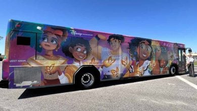 A city bus wrapped with colorful artwork featuring Disney’s "Encanto" characters, including Mirabel, butterflies, and vibrant patterns—a magical showcase of How to Get Around Disney World. A person stands near the front of the bus.