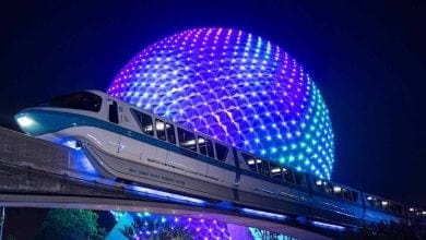 A futuristic monorail glides in front of the illuminated geodesic sphere of Epcot at night, glowing with vibrant blue and purple lights—a spectacular example of how to get around Disney World in style.