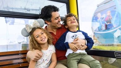 A smiling man sits on a gondola bench with two happy young children, one wearing Minnie Mouse ears. Outside the window, Disney-themed gondola cabins offer a glimpse of how to get around Disney World in style.