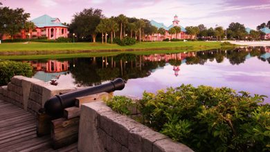 World Showcase lagoon at EPCOT, a cannon by lush gardens, pastel pavilions casting colorful sunset reflections in the water.