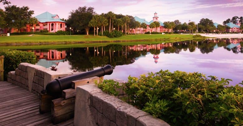 World Showcase lagoon at EPCOT, a cannon by lush gardens, pastel pavilions casting colorful sunset reflections in the water.