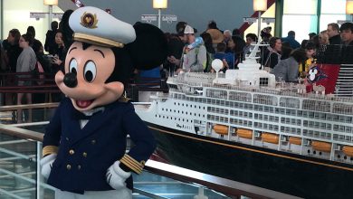 Mickey Mouse, dressed as a captain, greets guests by a Disney cruise ship display with excited fans gathered behind him.
