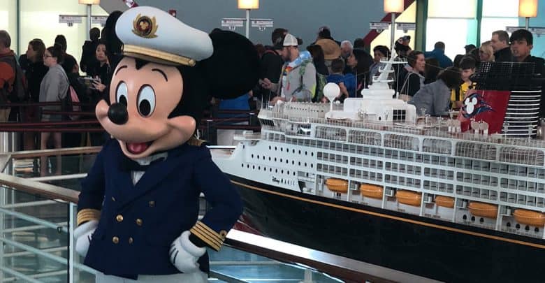 Mickey Mouse, dressed as a captain, greets guests by a Disney cruise ship display with excited fans gathered behind him.